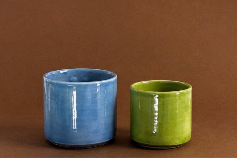 Two glazed ceramic tumblers in blue and green, displayed side-by-side against a solid brown background.